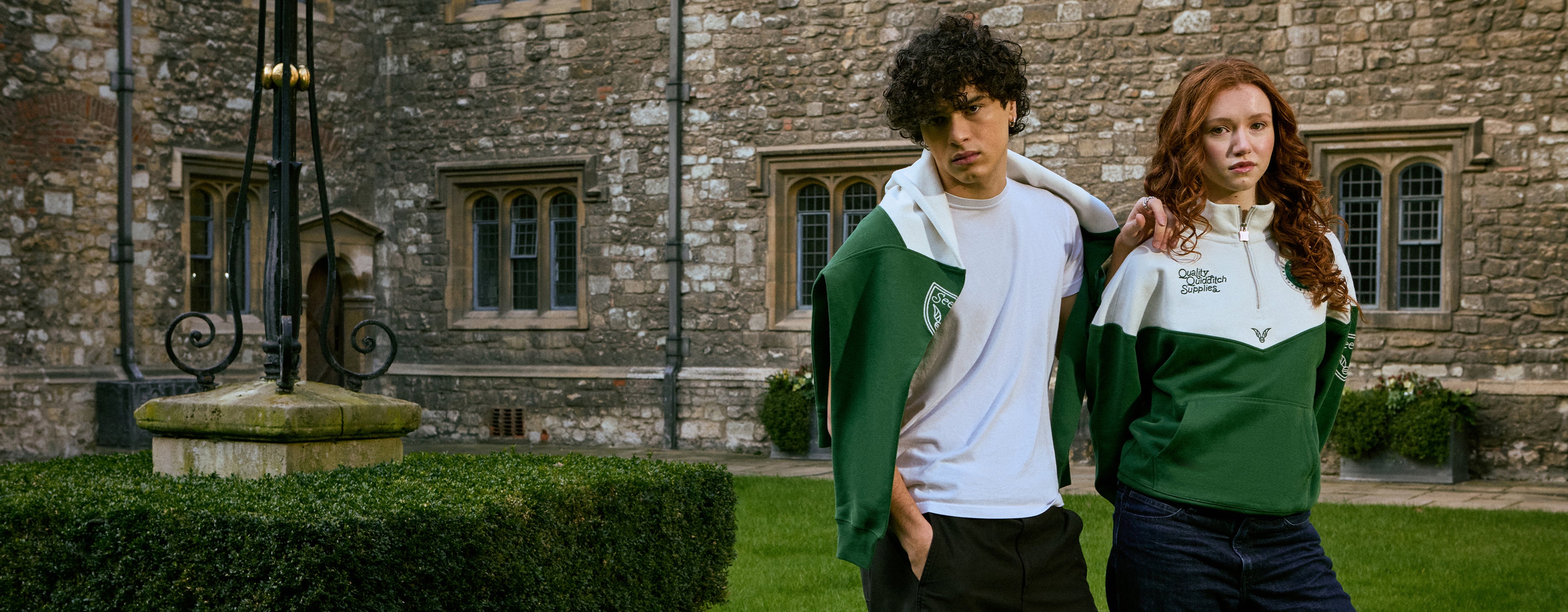 Two people wearing green and white Quidditch sweatshirts in front of a stone building.